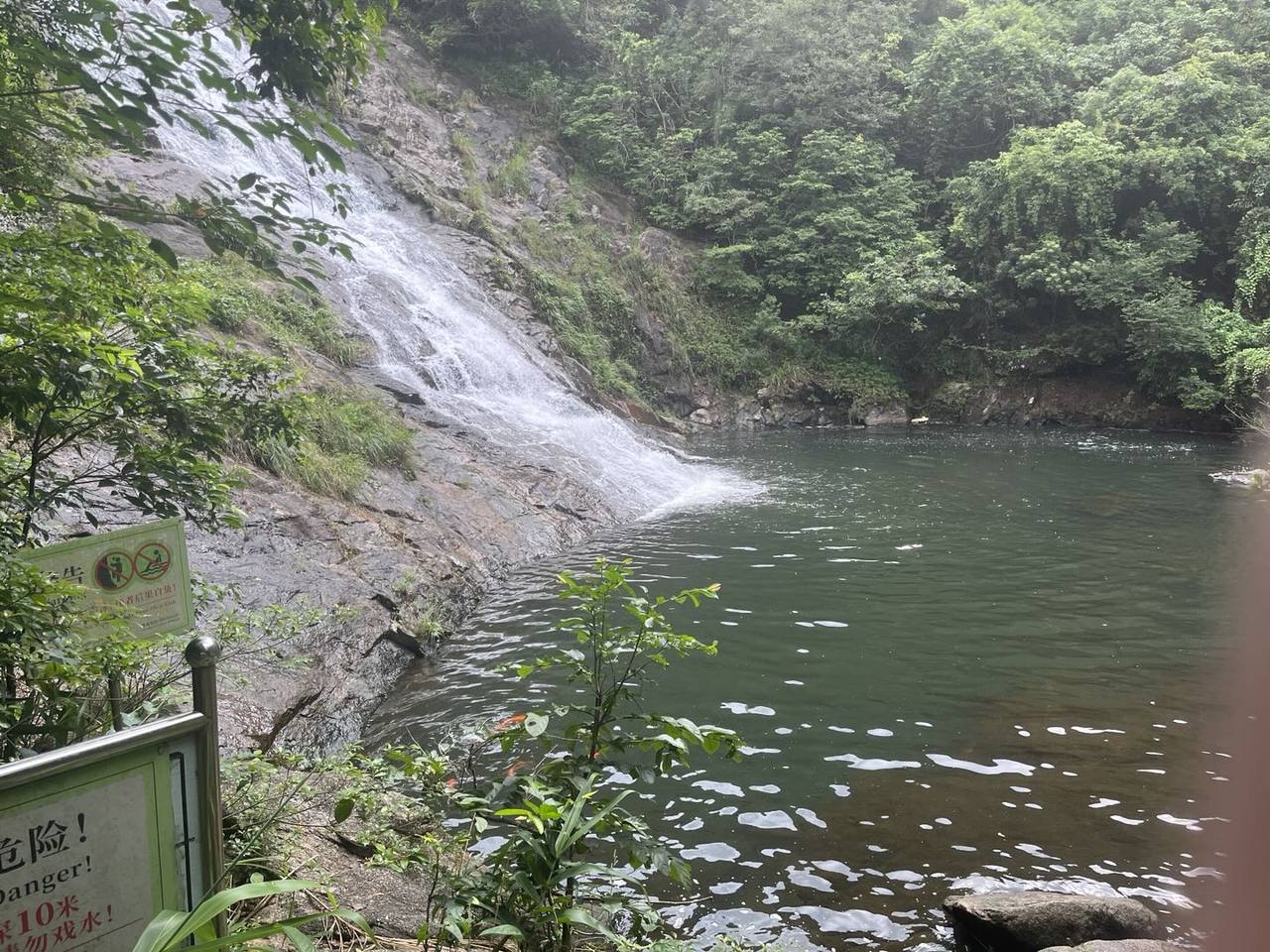 林间瀑布沿湿滑岩壁流入深绿水潭，前景有护栏和危险警示牌