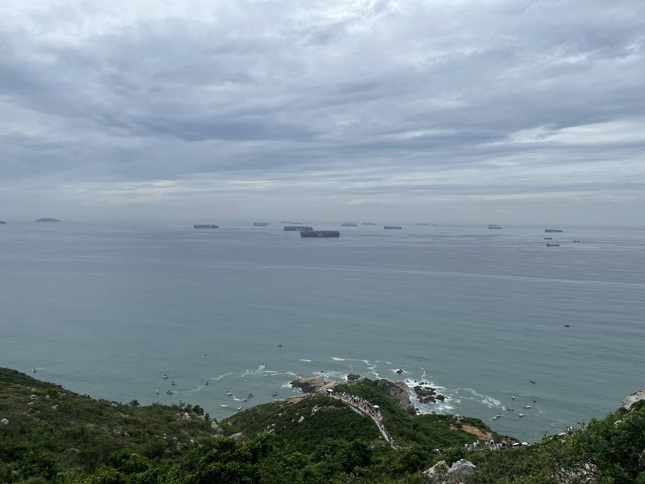 沿山脊步道俯瞰阴天海面,岩岸与礁石旁泛起白浪,远处海平线上可见多艘船只