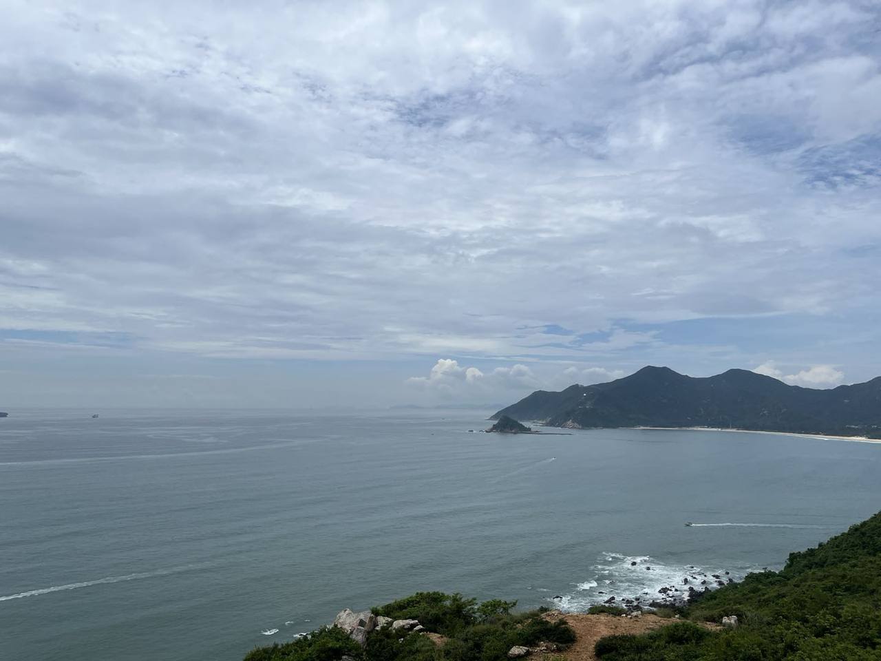 山峰处远望海天开阔,蓝灰海面延伸至云层下的天际,右侧深色远山半岛与浅色岸线相接;近岸礁石间浪花泛白,海上可见细小船影及几道淡淡航迹