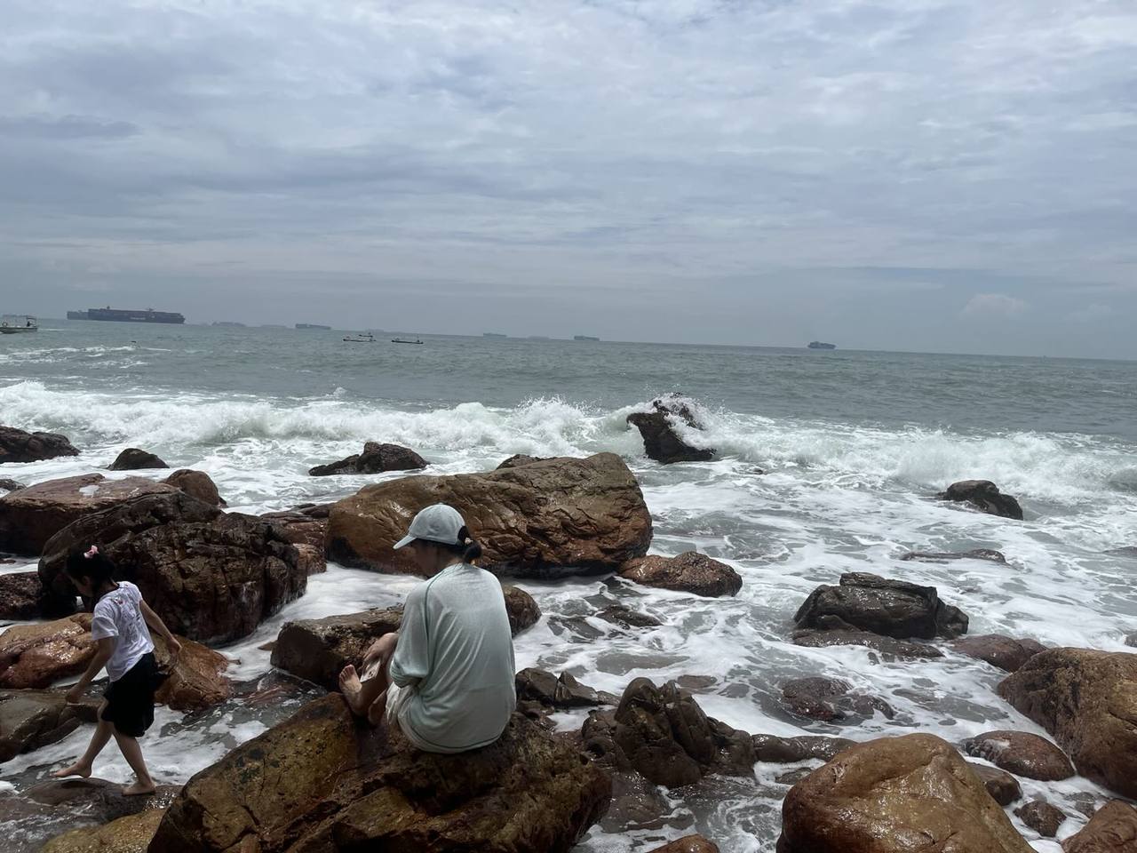 阴云下的礁石海岸,白色浪花在湿润的褐色岩石间翻涌,一人坐在近岸礁石上望向灰蓝色海面,远处可见零星船影