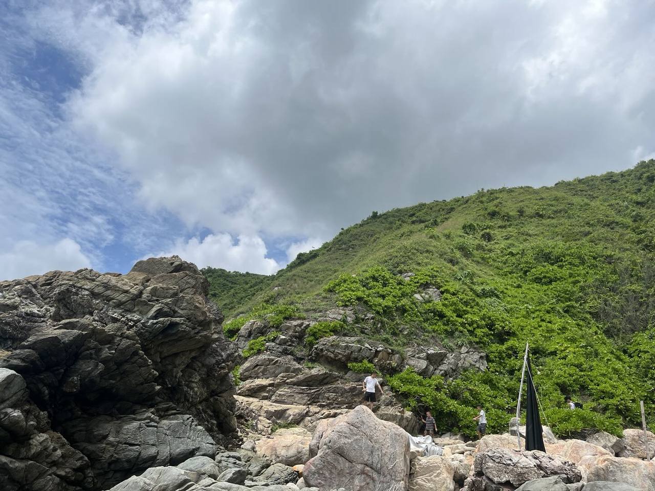多云天空下,绿植覆盖的山坡从右侧延伸至背景;前景散布着灰褐色巨石和层状岩壁,四名人物在崎岖岩石间行走或站立,右侧有一根挂着黑色布料的竖杆