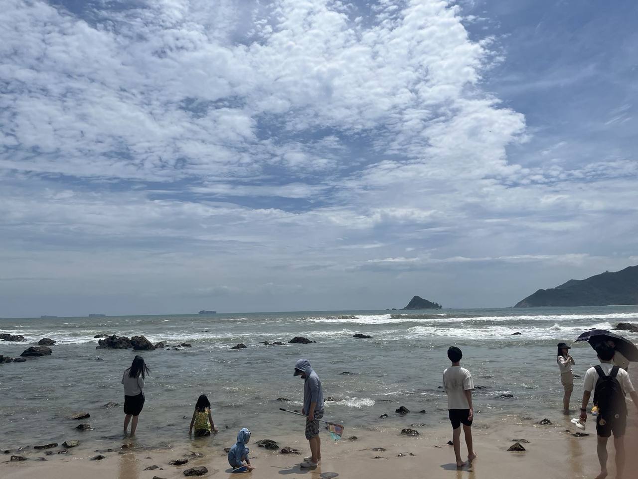 走出环线,踏进有礁石散布的湿沙与浅海;浪线越过近岸,远山和海平线上的船影映在云层下