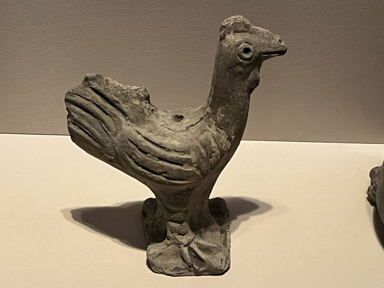 博物馆展柜中一件有磨损的灰褐色陶塑鸡形器
