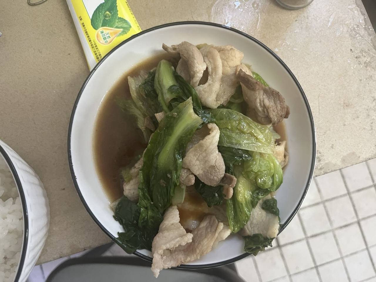 台面上的一碗绿色叶菜与浅色片状食物，配有棕色汤汁和白米饭。